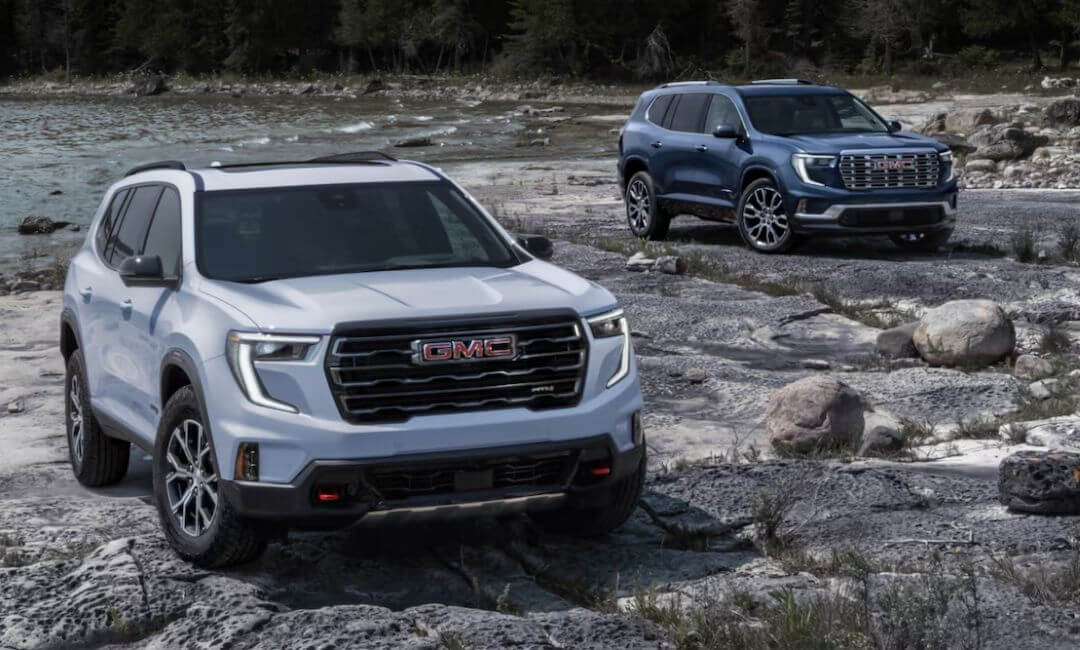 2025 GMC Acadia AT4 in white parked on a rocky lakeshore with a blue 2025 AcadiaDenali in the background